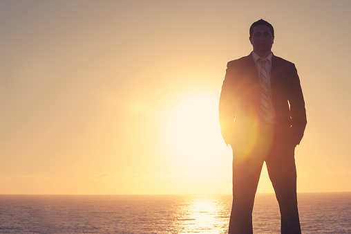 Successful Businessman standing confidently at sunrise. He is standing with his back to the sun as it rises behind him. This is a success, discovery, visionary concept. It could also portray confidence, power, authority, opportunity, exploration and innovation. Back lit silhouette style