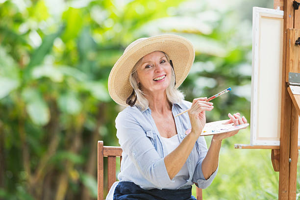Senior woman painting a picture on canvas A senior woman sitting in the park with canvas on an easel, painting a picture of the landscape.  She is holding a paint palette and a paintbrush, smiling at the camera.  It is a sunny day and she is surrounded by green foliage.  She is wearing a wide-brimmed hat and a light blue shirt, sitting on a wooden, folding chair. white hair photos stock pictures, royalty-free photos & images