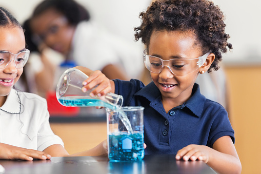 Adorable African American girl is student in private elementary school science class. She is pouring and combining blue chemicals during science experiment. Girl has natural curly hair and is wearing a blue private school uniform shirt. Classmate is watching excitedly.