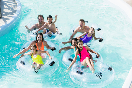 A group of eight people at a water park, floating on the lazy river on inflatable rings.  They are two families, friends having fun together on the water.  Each family has two children.  It is a high angle view.  They are smiling and waving.