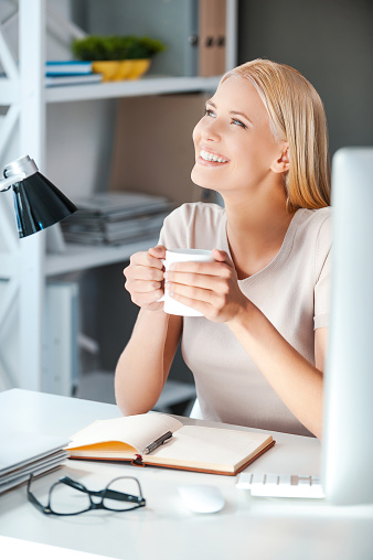 Beautiful young woman holding cup with hot drink and looking away with smile while sitting at her working place in office