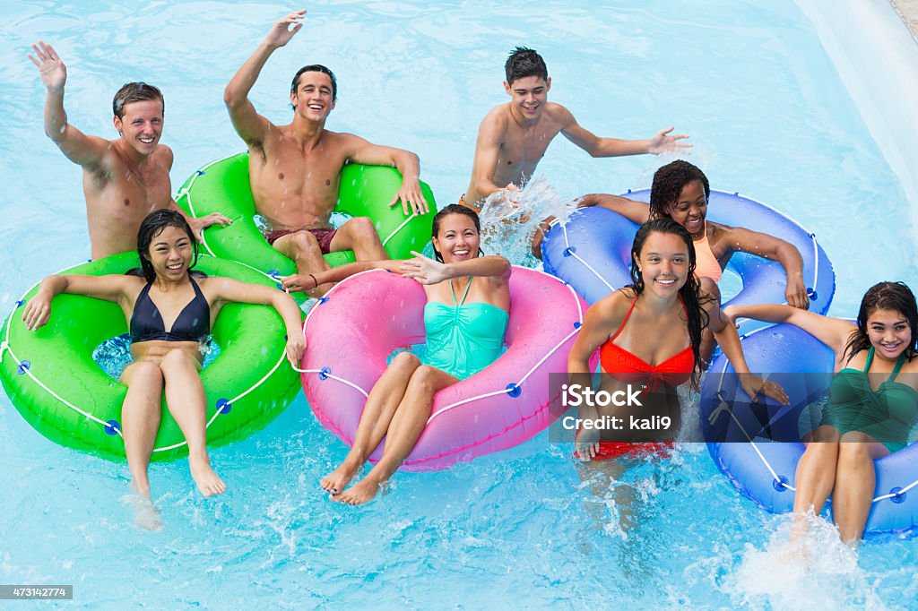 Teenage friends floating on innertubes A multi-ethnic group of teenagers hanging out at a water park, floating on the lazy river on colorful inflatable rings.  it is a high angle view of them waving at someone.  There are five teenage girls and three teenage boys wearing swimsuits in the swimming pool. African Ethnicity Stock Photo