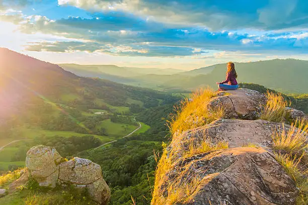 Woman doing yoga on top of a rock in the mountains, meditating in lotus posture, in a beautiful and colorful sunset. Energizing moment with the mother nature.