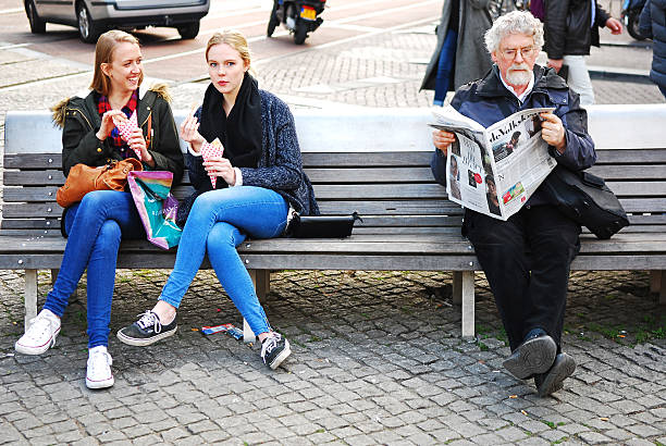 Amsterdam, The Netherlands - April 23, 2015: Two young women eating Potato Chips from a paper cone while sitting on a public bench in the center of Amsterdam. A man next to them reads a local newspaper.