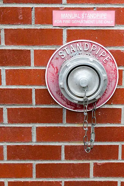 A metal manual standpipe is mounted in the exterior wall of a red, brick building.  The standpipe is used by the fire department in the case of a fire to run water through the building sprinkler system.  The single standpipe is weathered and isolated on a wall.