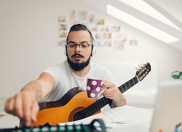 Musician Composing Music in his Recording Studio. Man with acoustic guitar composing music in his recording studio. Using laptop and writing lyrics or music notes. Wearing headphones and drinking coffee. Using sound mixer. acoustic guitar tattoos stock pictures, royalty-free photos & images
