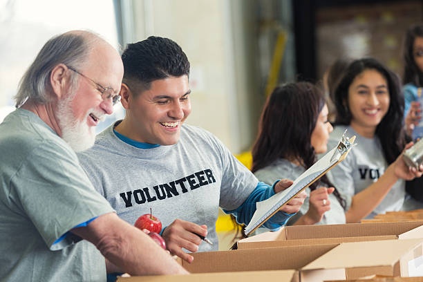 Senior man volunteering with younger adults in food bank Senior Caucasian man with grey hair and a beard is helping pack cardboard boxes with donated groceries in food bank. He is working with a Hispanic mid adult man who is wearing a VOLUNTEER t-shirt and holding a clipboard. Other volunteers are packing boxes with donations for less fortunate. word processing stock pictures, royalty-free photos & images