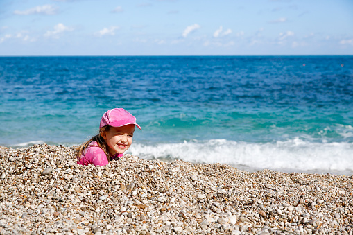 Girl buried in beach pebbles, smiling and enjoying free time on the beach, dressed in wetsuit and a hat for sun protection. Family and children on vacation, summer fun concept.
