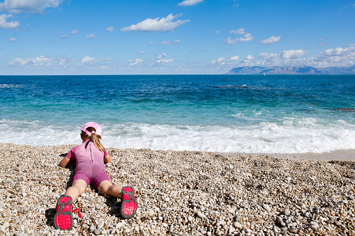 Girl lying on her belly, observing sea and enjoying free time on the beach, dressed in wetsuit and a hat for sun protection. Family and children on vacation, summer fun concept.