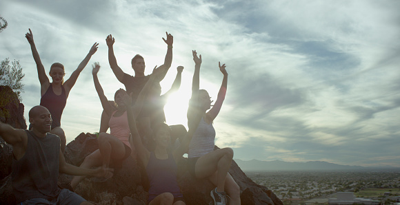 A multi-ethnic group of runners at the end of the trail on a mountain, looking out over the valley, their arms are raised in a cheer of victory.