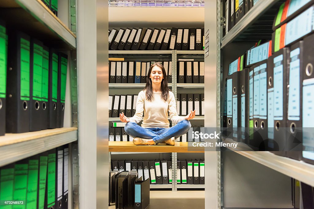 Woman meditating in company finance file archive Young woman meditating with closed eyes, taking a break on office desk in between the finance file archive, surrounded with ring binders, folders, files, papers in company document storage room. Wide angle concept shot. Made with Nikon D810. Archives Stock Photo