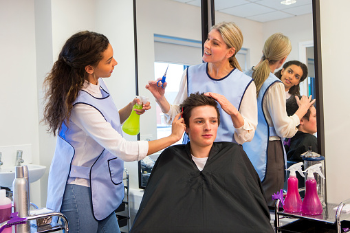 A horizontal image of a hairdresser and trainee hairdresser watching and learning. A young teenage boy sits in the chair receiving the treatment.