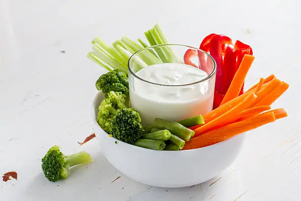 Vegetable sticks (pepper, celery, carrot, broccoli) in white bowl and yogurt, white wood background