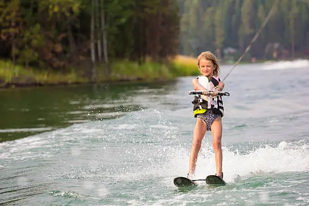 An action photo of a cute blonde little girl waterskiing beautiful scenic lake during summer vacation.