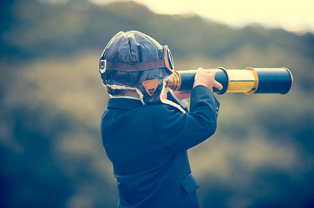Young boy in a business suit with telescope. Young boy in a business suit with telescope. Small child wearing a full suit and holding a telescope. He is holding the telescope up to his eye with an aviator cap on. Business forecasting, innovation, leadership and planning concept. Shot outdoors with trees and grass in the background projection stock pictures, royalty-free photos & images