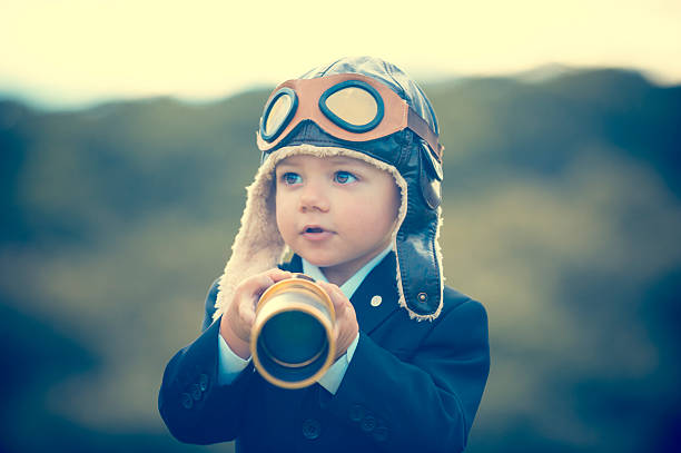 Young boy in a business suit with telescope. Small child wearing a full suit and holding a telescope. He is holding the telescope up to his eye with an aviator cap on. Business forecasting, innovation, leadership and planning concept. Shot outdoors with trees and grass in the background