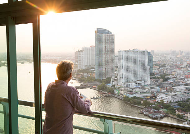 istock Man looks out from hotel railing, above city, river 473294566