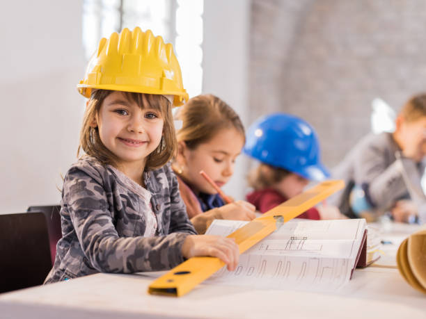 Little girl architect smiling and looking at camera. Happy little girl pretending to be an engineer and looking at camera. She is wearing yellow hard hat and holding level while sitting at the table in the office and working on construction plans. There are children in the background. career girl stock pictures, royalty-free photos & images