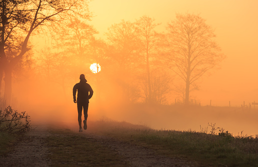 Athlete running on a gravel road during a foggy, spring sunrise in the countryside, with the sun in the background.