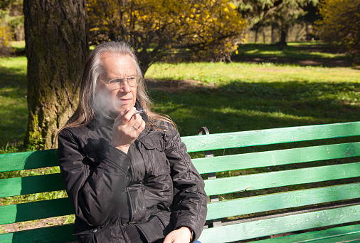 elderly gray-haired man in glasses, black jacket smoking a cigarette sitting on a bench in city park on sunny spring day