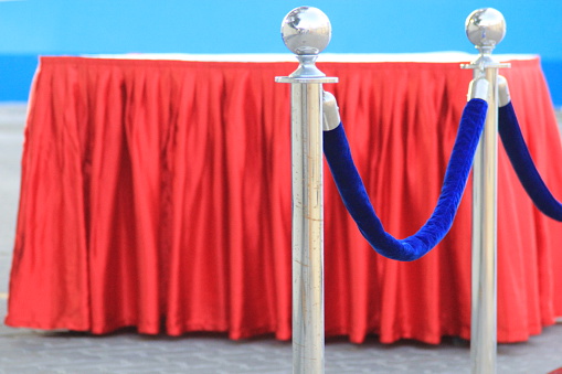 Crowd control barriers with blue velvet rope with a table covered in red fringe ready to accept guests to a function, party, reception or celebration