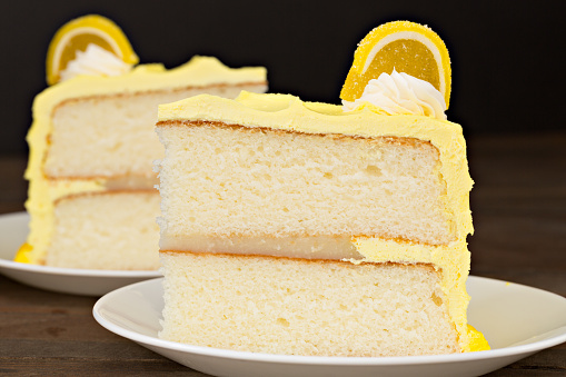 A close up horizontal photograph of two slices of layered lemon cake, the layers are separated by a lemon filling and the creamy lemony frosting is topped with some white whipped cream and a jelly lemon slice.