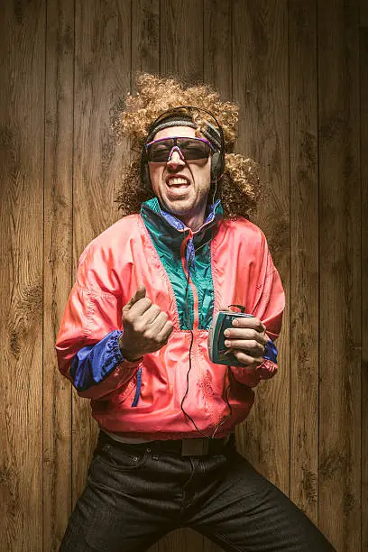 A funky man in late 1980's / early 1990's fashion style, with curly hair and fluorescent colored track suit listening to a walkman tape player and wearing ear muff headphones.  He sings out loud to the song,  dancing to the music.  Wood paneling in the background.  Vertical portrait.