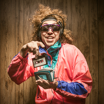 A funky hipster man in late 1980's / early 1990's fashion style, with curly hair and fluorescent colored track suit listening to a walkman tape player and wearing ear muff headphones.  He smiles with a cheesy grin, putting a cassette tape into the player with great excitement.  Wood paneling in the background.  Square crop.