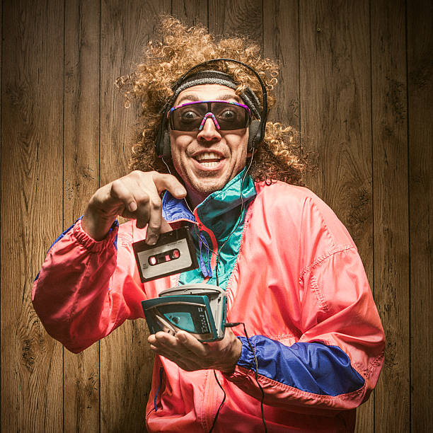 Fashion of Nineteen Eighties and Nineties with Walkman A funky hipster man in late 1980's / early 1990's fashion style, with curly hair and fluorescent colored track suit listening to a walkman tape player and wearing ear muff headphones.  He smiles with a cheesy grin, putting a cassette tape into the player with great excitement.  Wood paneling in the background.  Square crop. personal stereo stock pictures, royalty-free photos & images