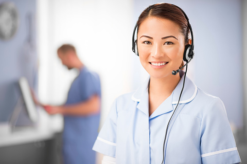 a young female nurse talks into a headset . She is wearing medical scrubs and looks to camera chatting on the phone. In the background we can see a blurred clinic setting with another nurse defocussed .