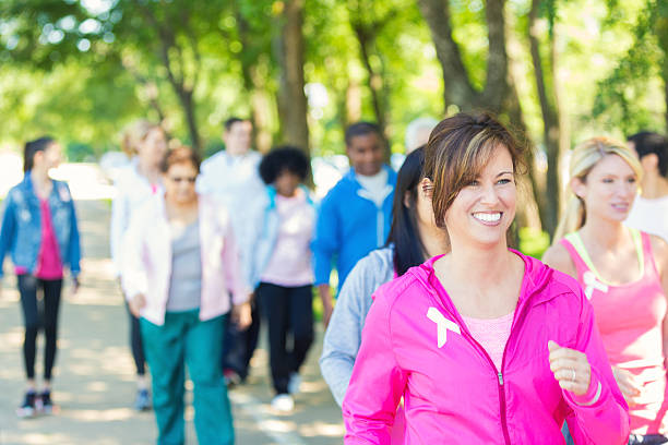 Woman walking in breast cancer awareness charity race event Mature Caucasain brunette woman is smiling while walking in charity marathon race event. She is wearing pink athletic clothing and a breast cancer awareness ribbon. Diverse teams of people are walking behind her to raise money for breast cancer research. womens issues stock pictures, royalty-free photos & images