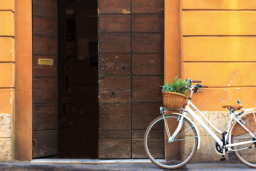 Charming and peaceful Roman scene: white bike with green herbs in the basket resting against a vibrant yellow wall and big brown doors. Copy space available on the doors or wall.