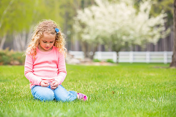 Front view of a young girl with curly blonde hair sitting in the grass on a springtime day. A tree full of white blossoms can be seen in the distance. The girl is looking down at something she is holding in her hand.
