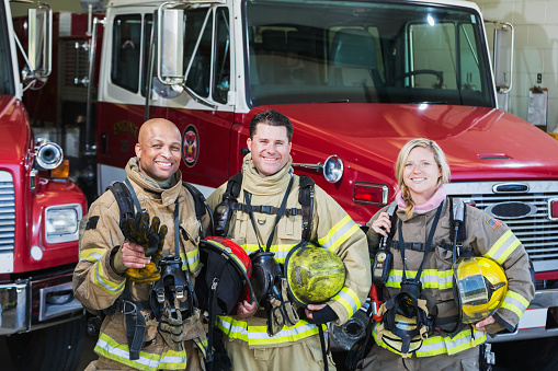 Portrait of a diverse group of three firefighters at the station standing in front of fire trucks with their protective gear.  They are wearing tan suits with yellow reflective stripes, holding helmets and protective gloves.