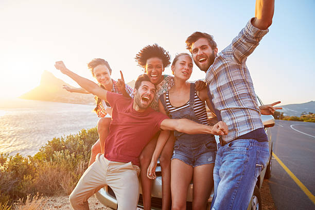 Friends standing by a car next to a coastal road at sunset Group Of Friends Standing By Car On Coastal Road At Sunset couple driving at night stock pictures, royalty-free photos & images