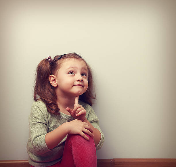 Fun thinking kid girl sitting in pink jeans and looking up on empty copy space. Vintage portrait