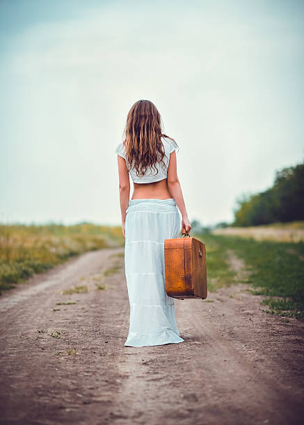 istock Young woman with suitcase going away by field road 473458742