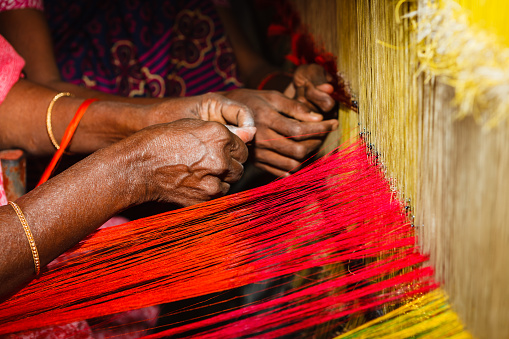Kanchipuram, India - January 29, 2015: A couple of women load silk thread into a hand-loom, weaving the famous, much sought after, Indian Conjeevaram or Kanchipuram silk sari in the town of Kanchipuram in Tamil Nadu, South India. This is a centuries old craft, that flourishes in this one town in the whole of India.  Kanchipuram saris are a must at South Indian Tamil weddings. Photo shot in the horizontal format.  Focus on the red threads and hand in foreground.