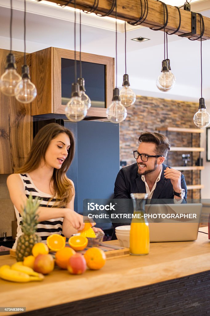 Young couple in the kitchen Young woman is squeezing orange while her male friend is working on laptop computer. They look like starting their morning together before leaving for work. On the table are oranges, apples, bananas, pineapple, bottle of orange juice and a toaster. 2015 Stock Photo