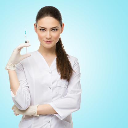 Beautiful young doctor in medical robe holding syringe with liquid. Isolated over blue background,