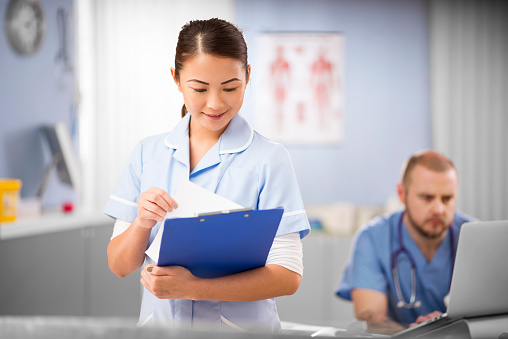 A young women nurse wearing blue medical scrubs and a stethoscope stands looking at her medical notes. In the background a hospital interior is shown with a defocussed male doctor sitting at his desk and working at a computer .