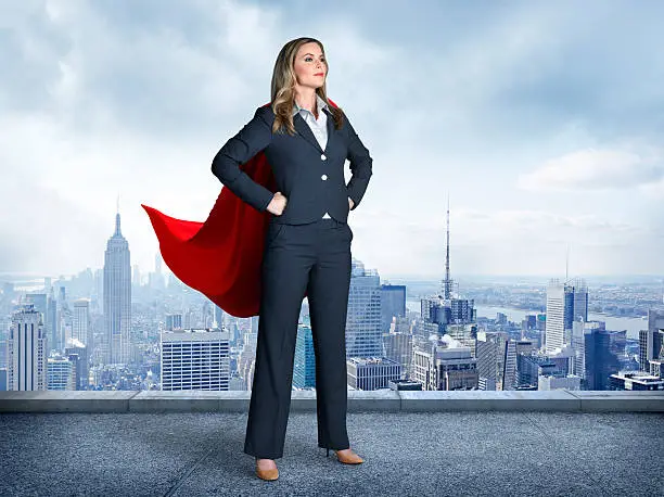 A businesswoman wearing a red cape strikes a super hero pose.  Her  hands on her hips and her feet are apart as she stands in front of the New York skyline.