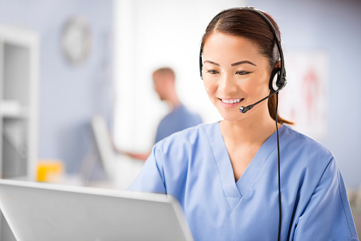 a young female nurse talks into a headset . She is wearing medical scrubs and looks to camera chatting on the phone. In the background we can see a blurred clinic setting with another nurse defocussed .