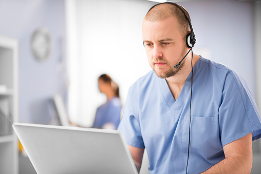 a male nurse talks into a headset . He is wearing medical scrubs and looks at his computer whilst chatting on the phone. In the background we can see a blurred clinic setting with another nurse defocussed .