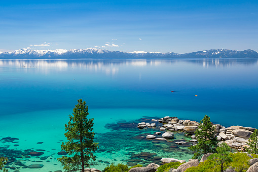 Picture of Lake Tahoe from east shore. There is a snow on the Sierra Nevada Mountains and some white clouds with reflection in turquoise waters of the lake. There are two kayaks on the calm waters. 