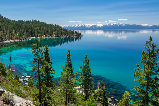 Picture of Lake Tahoe from east shore. There is a snow on the Sierra Nevada Mountains and some white clouds with reflection in turquoise waters of the lake. 