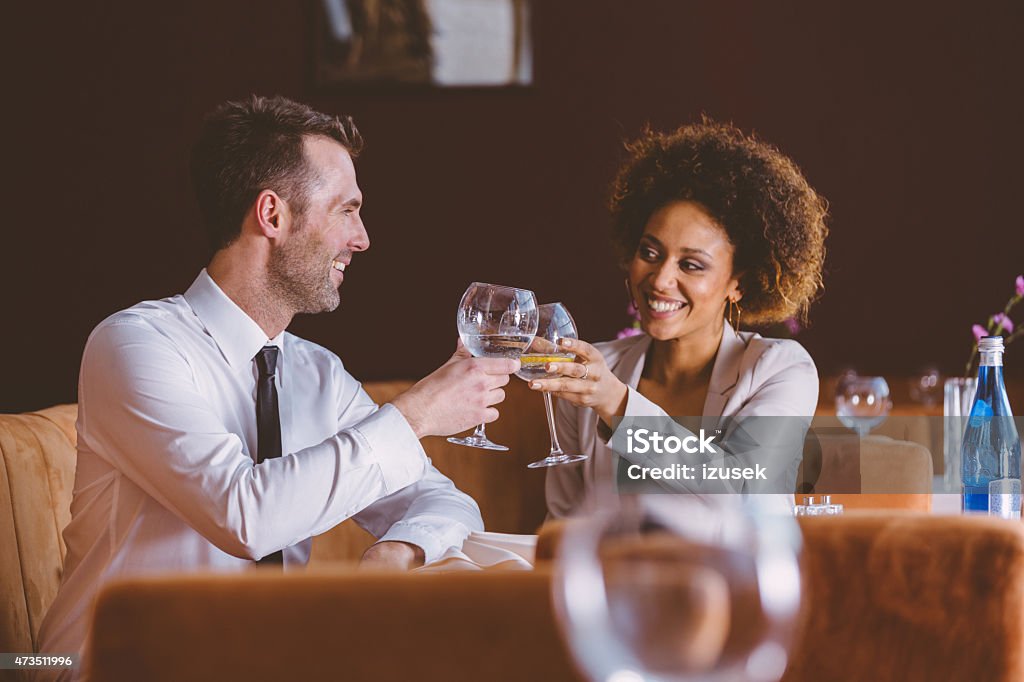 Two happy business people at lunch in the restaurant An afro american businesswoman and caucasian businessman having lunch or dinner in restaurant, sitting at the table, laughing and toasting with glass wine. African Ethnicity Stock Photo