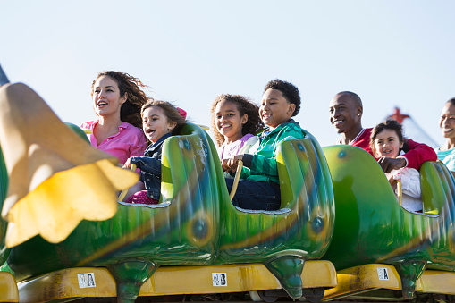 A group of seven multiracial people, children and adults, riding a rollercoaster at an amusement park.