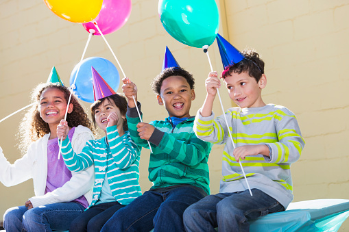 A group of four multi-ethnic children at a birthday party.  They are sitting in a row wearing party hats and holding colorful balloons.  The African American boy and girl are smiling at the camera.  They are wearing jeans and lond-sleeved, striped shirts.
