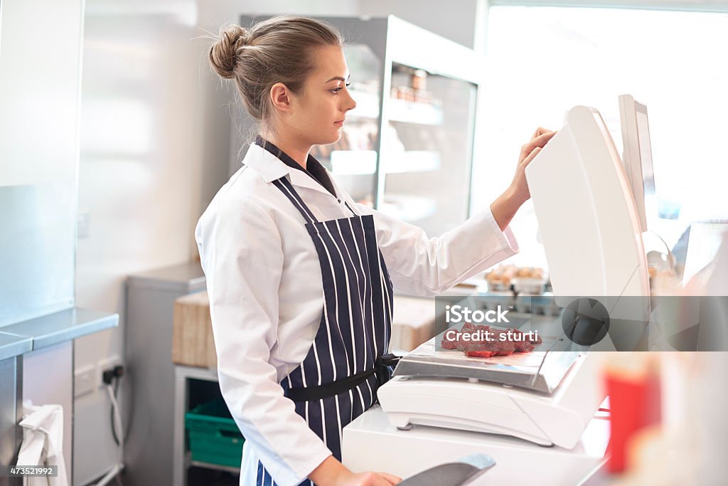 young female butcher portrait a young female butcher tends the meat counter in a butcher's shop. She is wearing a white coat and striped apron and is using a touchscreen weighing device. She is using electronic scales to weigh some diced steak . 20-29 Years Stock Photo